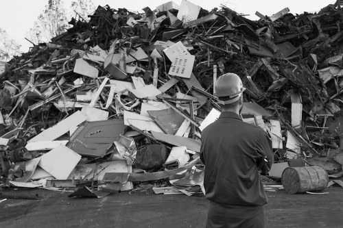 A man wearing a safety helmet and dark work clothing stands with his back to the camera, observing a large, chaotic heap of mixed construction and household waste materials on an outdoor asphalt surface. The rubbish includes broken and discarded items such as plastic containers, metal scraps, wooden pieces, cardboard, and other debris, all piled high and appearing to be in various stages of disassembly. The textures range from smooth plastic surfaces to rough wood and metallic edges, with some items stacked and others scattered in a disorganized manner. In the background, there is a clear sky and a few distant trees, indicating the rubbish pile is situated on a driveway or industrial yard, possibly in the vicinity of Abbots Langley. The scene reflects a waste management or clearance operation, where firms like House Clearance Abbots Langley might handle the removal of such bulky debris efficiently from residential or commercial properties.