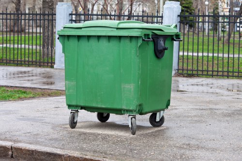 A large green plastic wheeled rubbish bin with a closed lid, situated on a paved surface outdoors. The bin features a textured surface with vertical ridges along its sides, a black wheelie ban handle on the top front edge, and four black caster wheels at the base for mobility. It is positioned in front of a black metal fence with decorative vertical bars, behind which there are trees with bare branches and a grassy area, suggesting a park or communal outdoor space. The scene appears to be during damp weather, as the pavement shows signs of moisture, and the overall environment is clean and orderly, suitable for waste collection or rubbish removal services in the local area such as Abbots Langley.