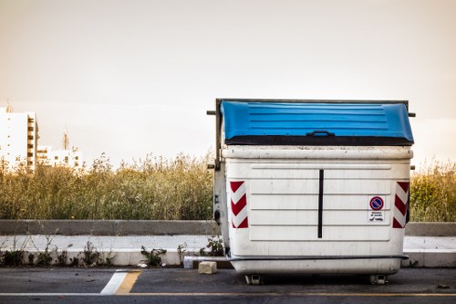 A large outdoor metal rubbish skip with a blue plastic lid positioned on a paved surface, likely a car park or service area. The skip is predominantly white with vertical ridges on its sides and features red and white reflective safety markings along its edges for visibility. There is a small circular no-parking sign attached to the front of the skip, and the skip's lid is closed, with a slight horizontal opening in the middle. In the background, a strip of overgrown grass and shrubs separates the paved area from a distant view of bare-branched trees and a low horizon, under a pale, overcast sky. The environment suggests a setting suitable for waste collection and disposal, potentially related to a local rubbish removal service like House Clearance Abbots Langley, situated near the outskirts of town, possibly in a commercial or industrial zone.
