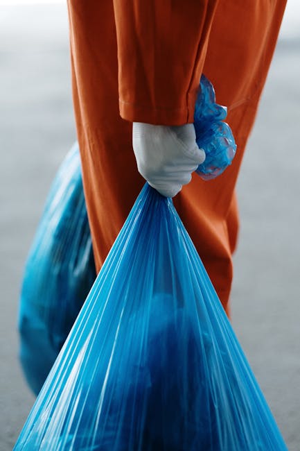 A person wearing a yellow and grey checkered shirt, grey trousers, and bright green gloves is holding open a large black rubbish bag lined with a silver inner layer, standing outdoors on a grassy area