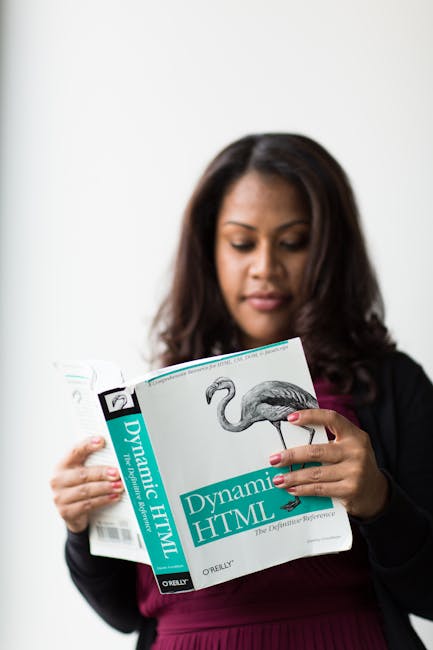 A woman with dark, shoulder-length hair is holding a book titled 'Dynamic HTML' published by O'Reilly, which features a cover illustration of a flamingo. She is standing indoors against a plain, light