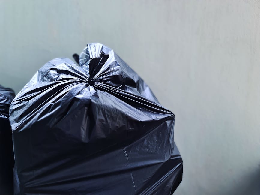 A black wheeled rubbish bin labeled 'St. John's' is positioned on the edge of a sidewalk on a quiet urban street at night. The bin is filled with miscellaneous waste, including cardboard boxes, paper,